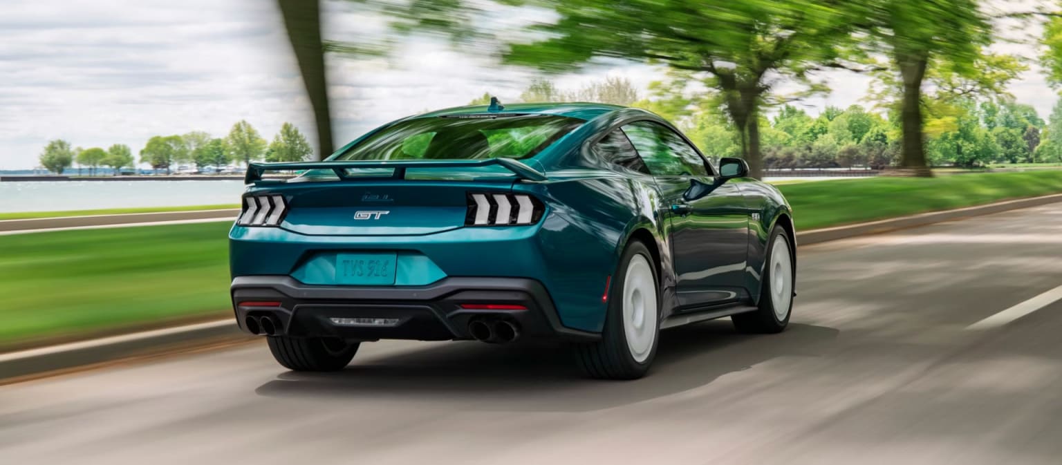 A sleek, teal-colored sports car speeds down a road surrounded by lush, green trees and a blurred background, conveying a sense of power and motion.
