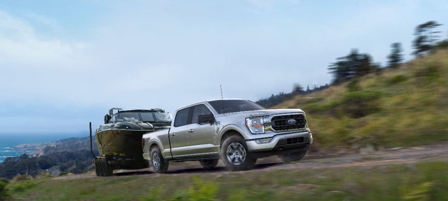 a silver F150 truck pulling a trailer on a dirt road next to a hill with a view of the ocean