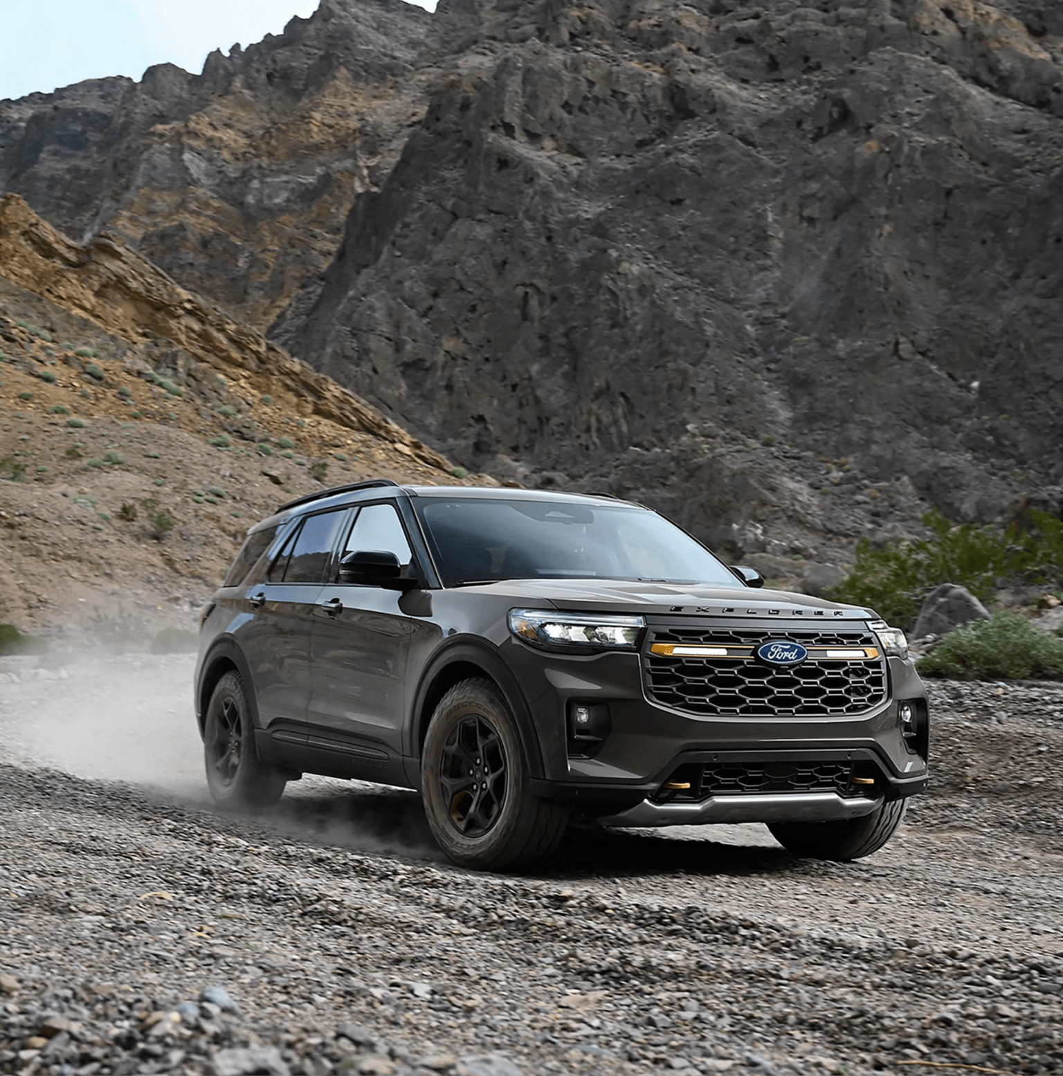 A grey SUV drives across a rocky dirt path with towering, rugged mountains in the background.