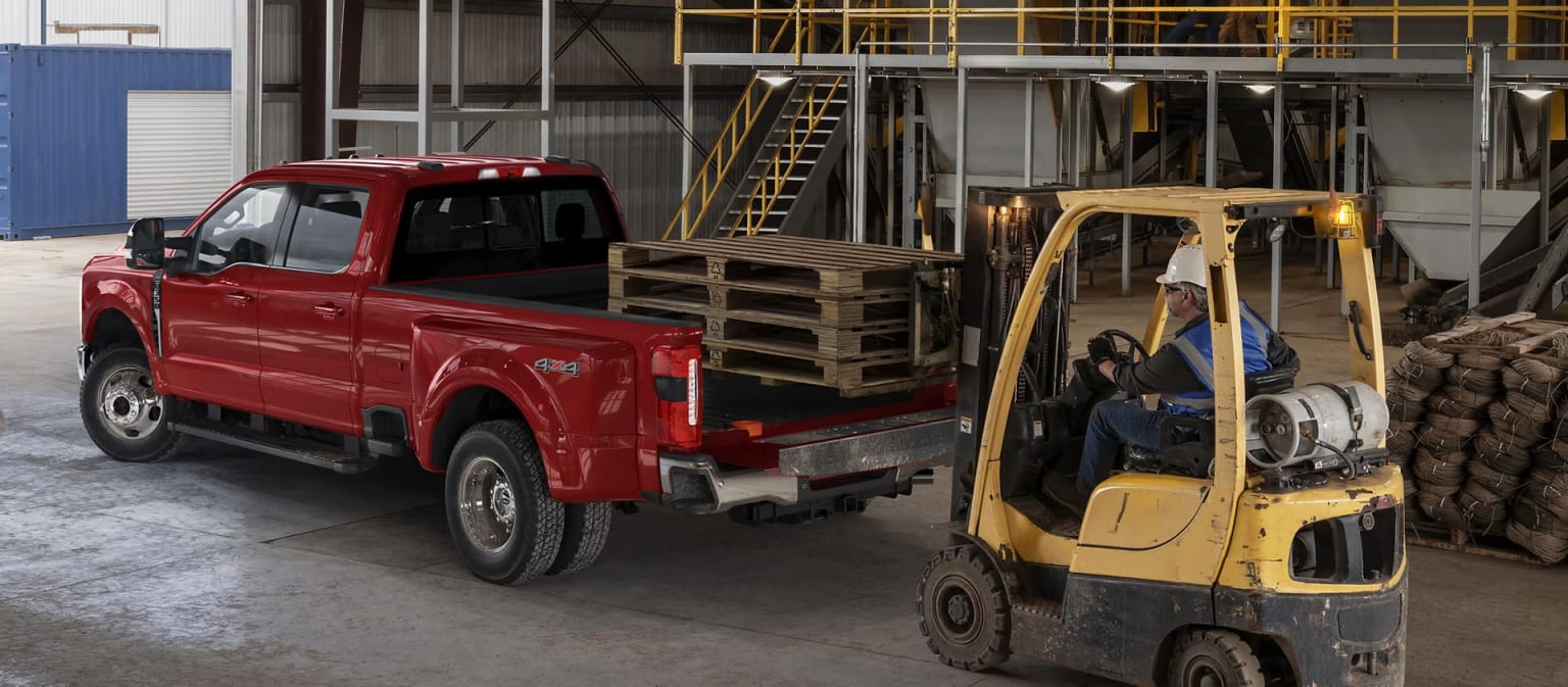 A red pickup truck in the foreground, with a forklift and various equipment and materials in the background, all situated in an industrial warehouse setting.