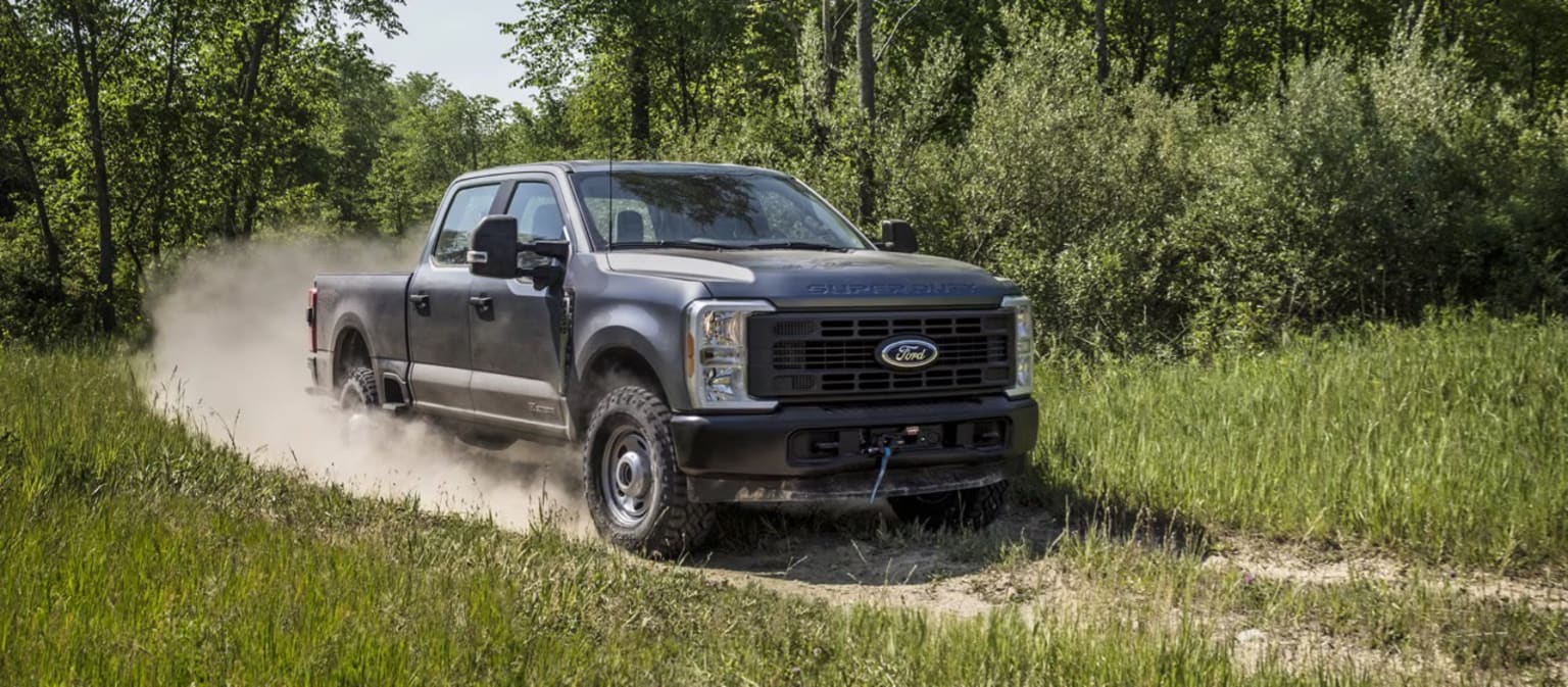 Off-road pickup truck driving through a grassy field, kicking up dust as it navigates uneven terrain surrounded by lush, green foliage.