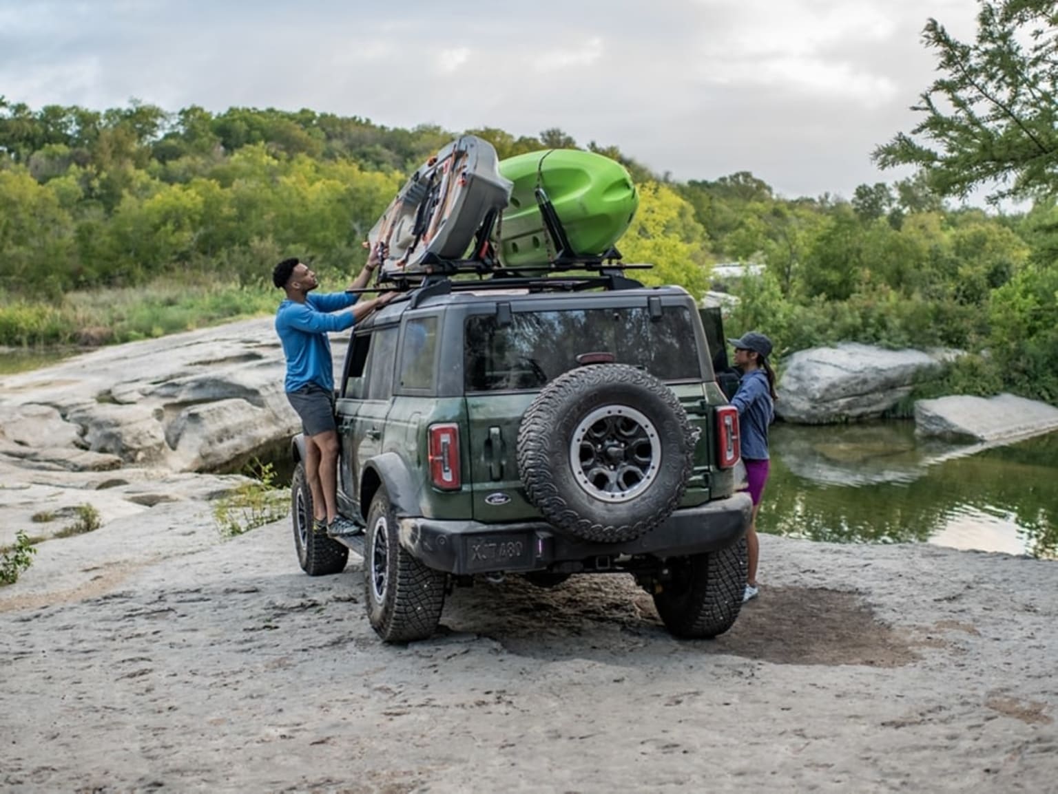 An off-road vehicle parked on a rocky riverbank surrounded by lush greenery and a tranquil body of water, with various outdoor gear and equipment.