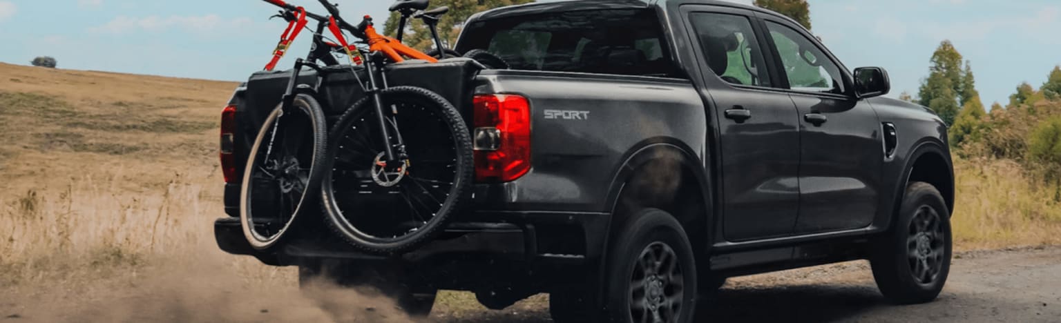 A dark gray pickup truck with two bicycles in the bed kicks up dust on a dirt road with a grassy hill and trees in the background.