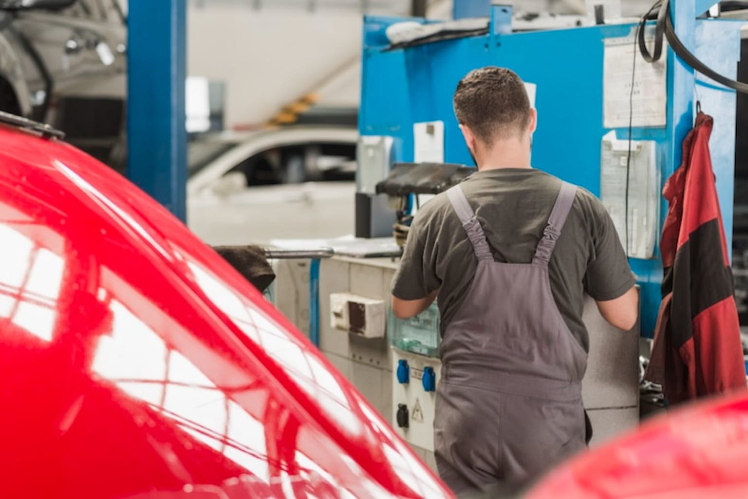 A mechanic in a gray jumpsuit stands in a workshop surrounded by various tools and equipment, with a bright red car in the foreground.