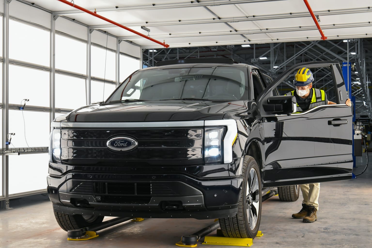 a man standing next to a black truck in a garage next to another man in a yellow safety helmet