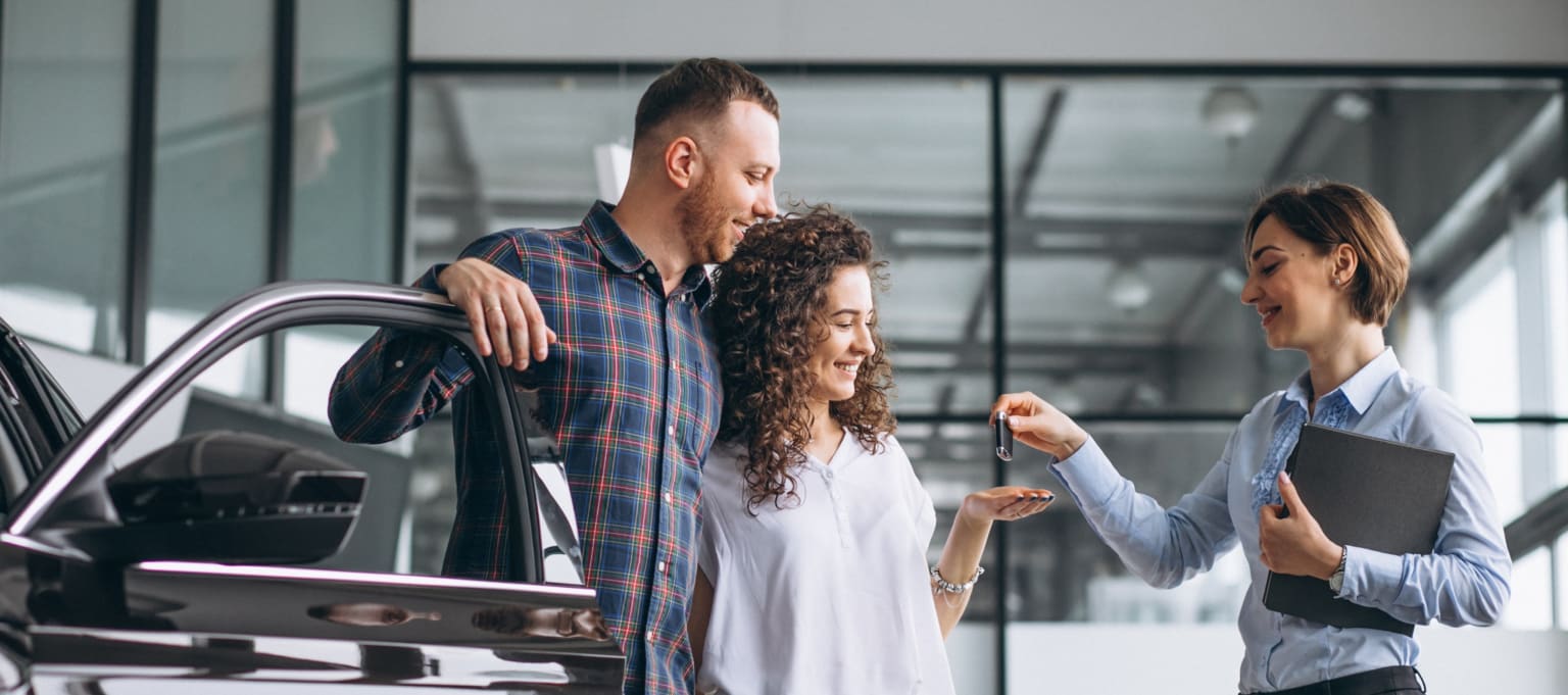 A couple receives car keys from a salesperson in a showroom with large windows.