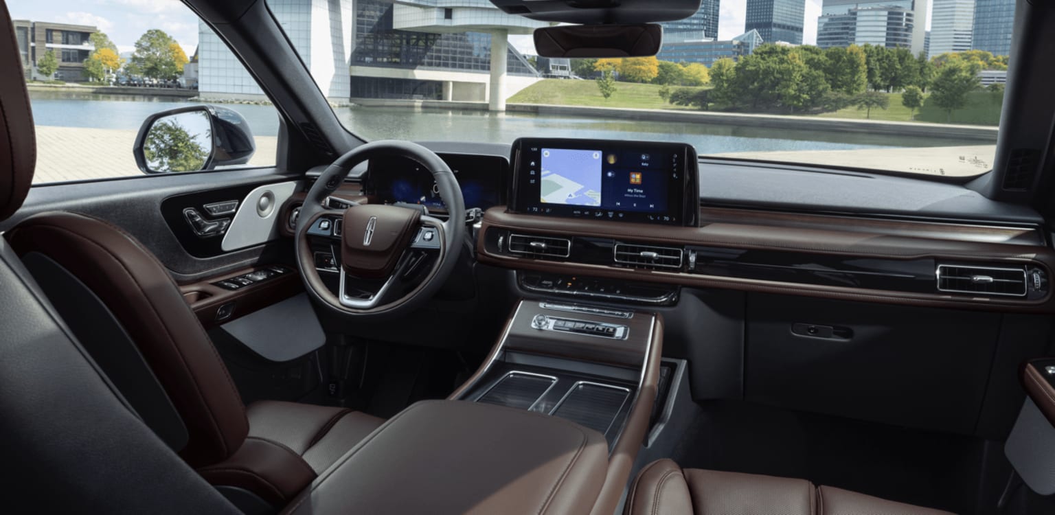 The image shows the interior of a modern vehicle with a sleek dashboard, a large touchscreen display, and a steering wheel with various controls. The background outside the vehicle appears to be a cityscape with tall buildings and greenery.