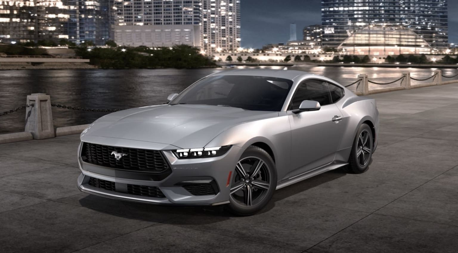 Silver sports car parked on a riverfront promenade with skyscrapers in the background at night.