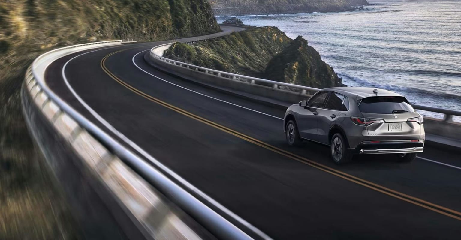 A silver SUV driving along a winding coastal highway with the ocean on the right and a blurred hillside on the left.