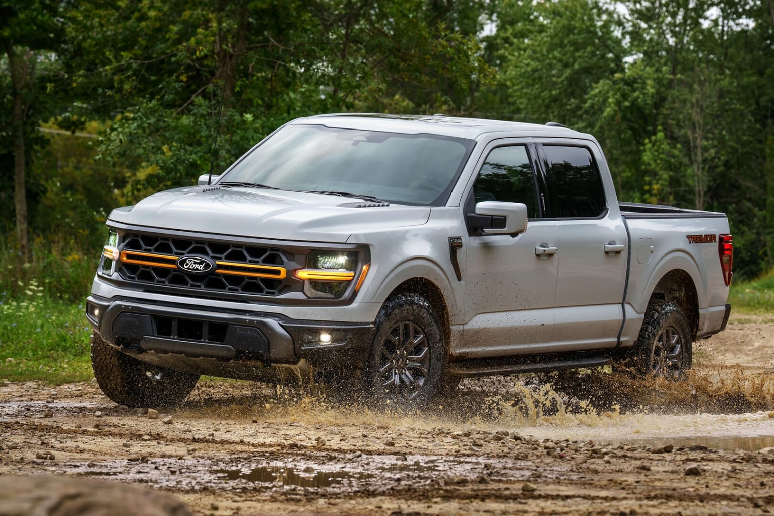 The image shows a silver pickup truck driving through a muddy, wooded area with lush green foliage in the background.
