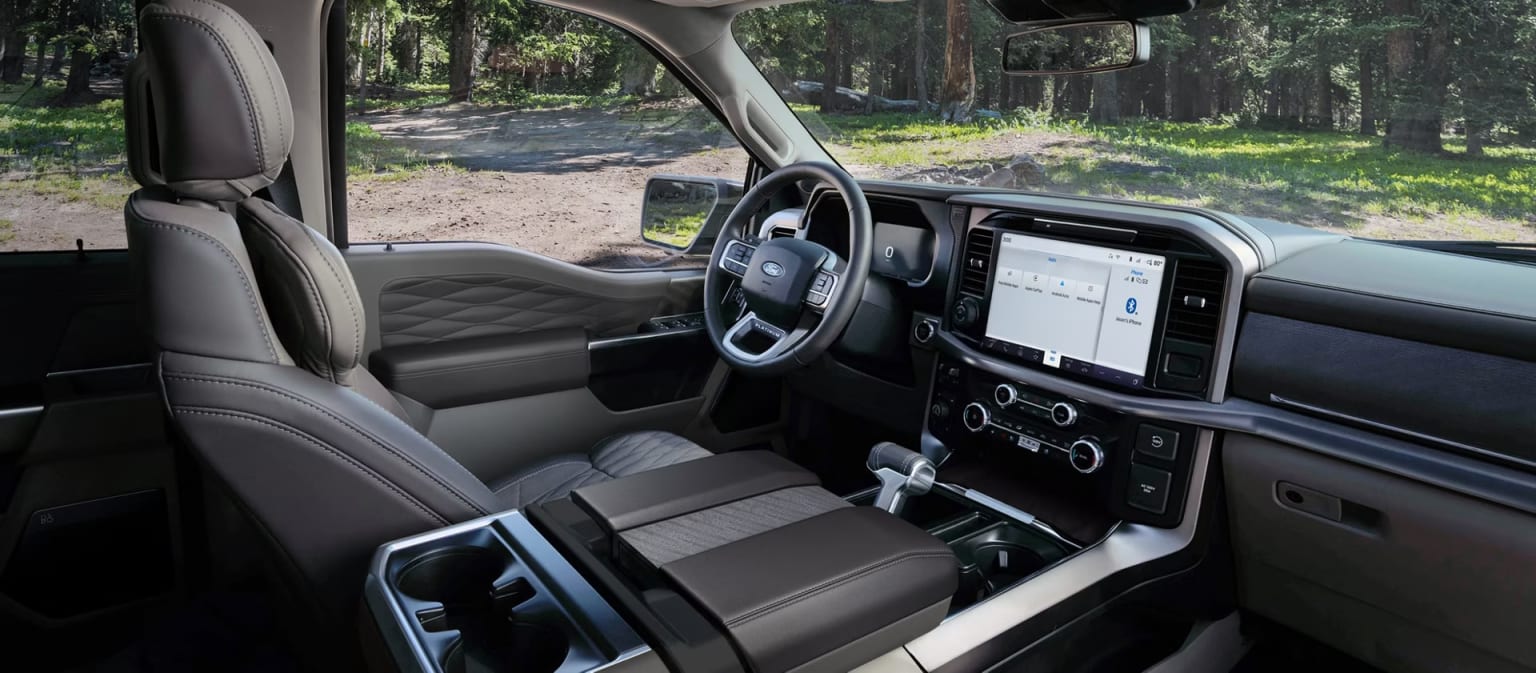 Luxurious interior of a truck with leather seats and a large touchscreen, set against a backdrop of a wooded area with trees and a dirt road.