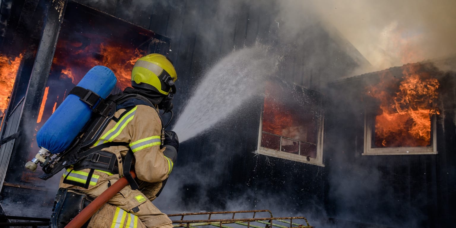 A firefighter in protective gear is using a high-pressure water hose to extinguish a raging fire in a building, with flames and smoke visible in the background.