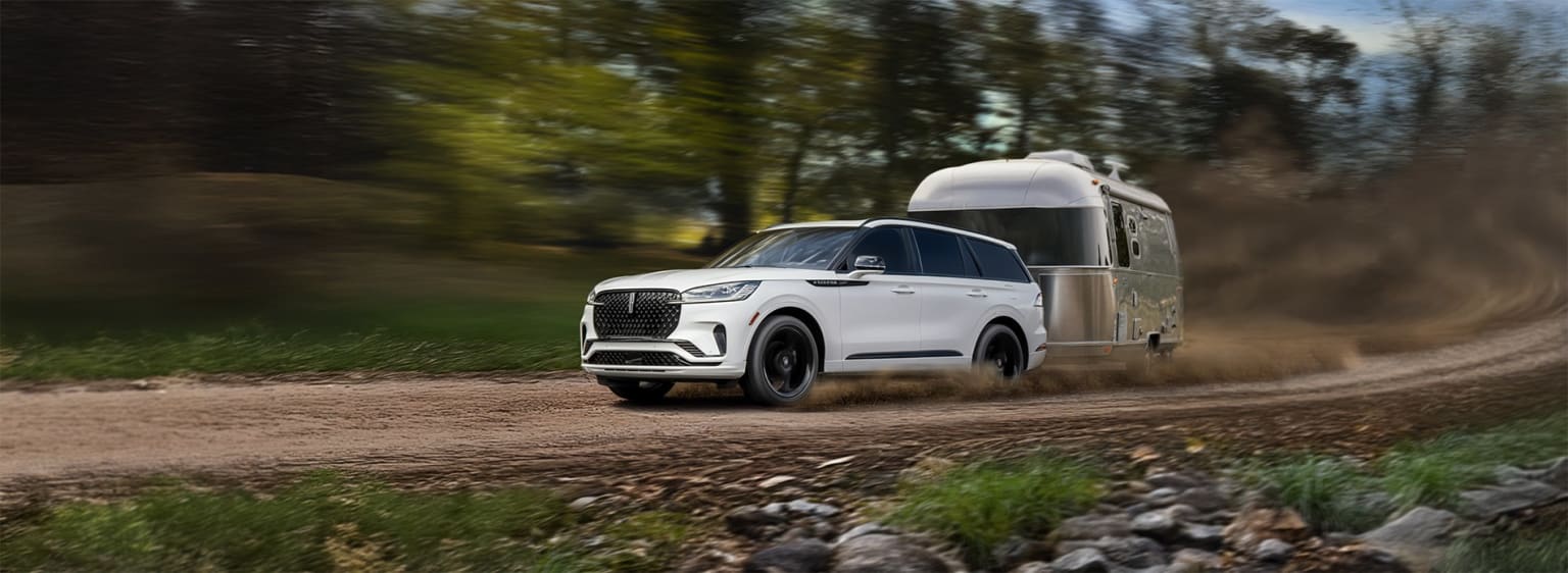 A large white SUV with a trailer attached driving down a dirt road surrounded by lush greenery and trees.