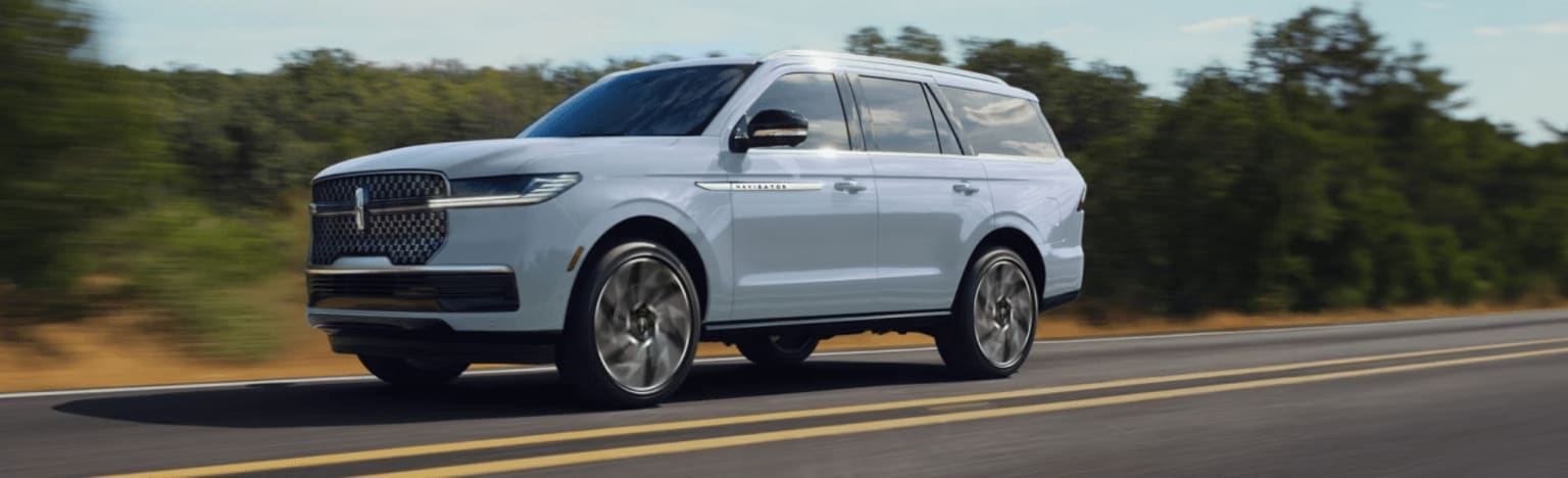 A light blue SUV drives down a paved road with trees lining the background.
