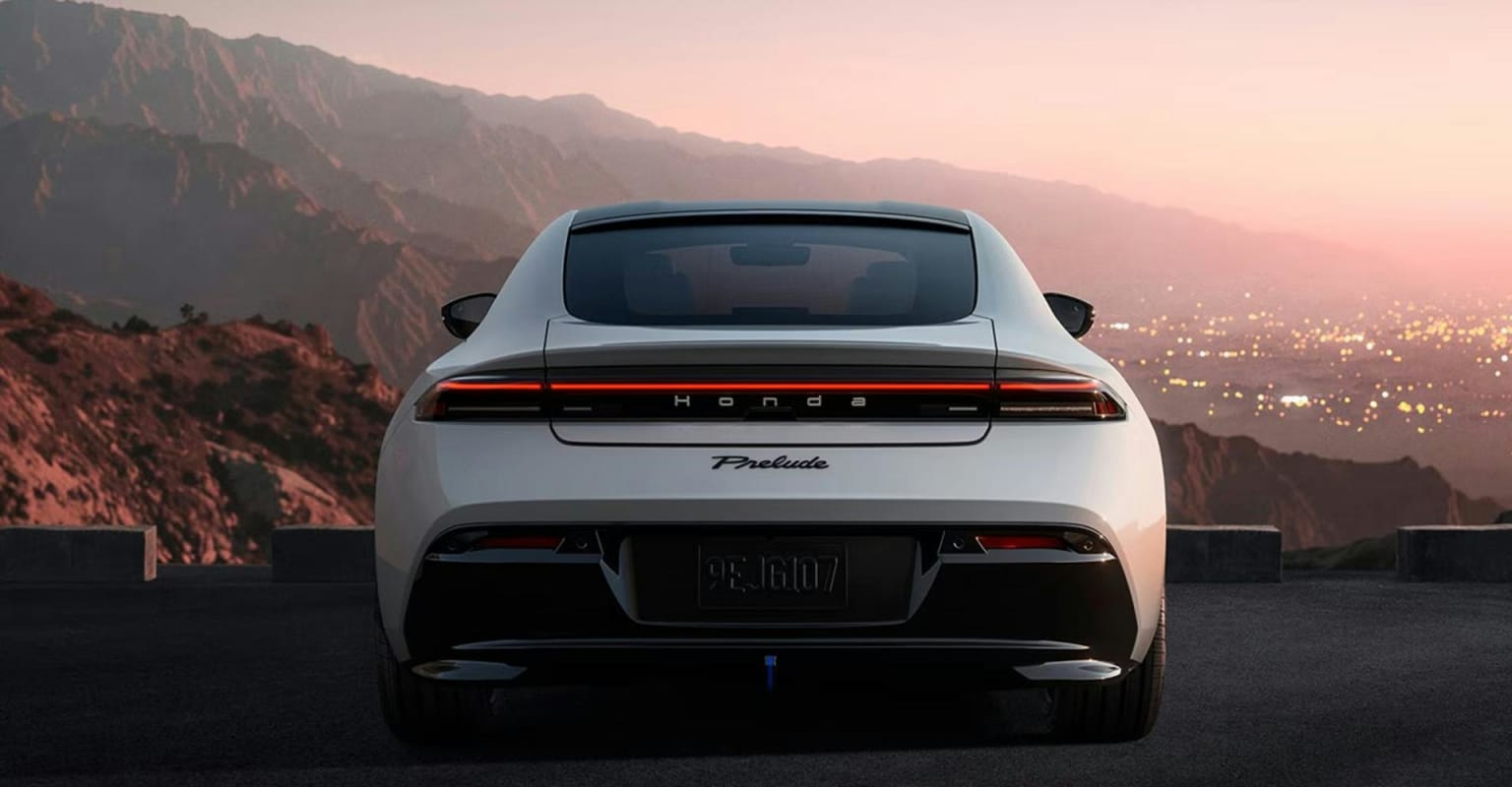 A white electric car with a sleek rear design parked on a road overlooking a sprawling city at dusk, with mountains in the distance.