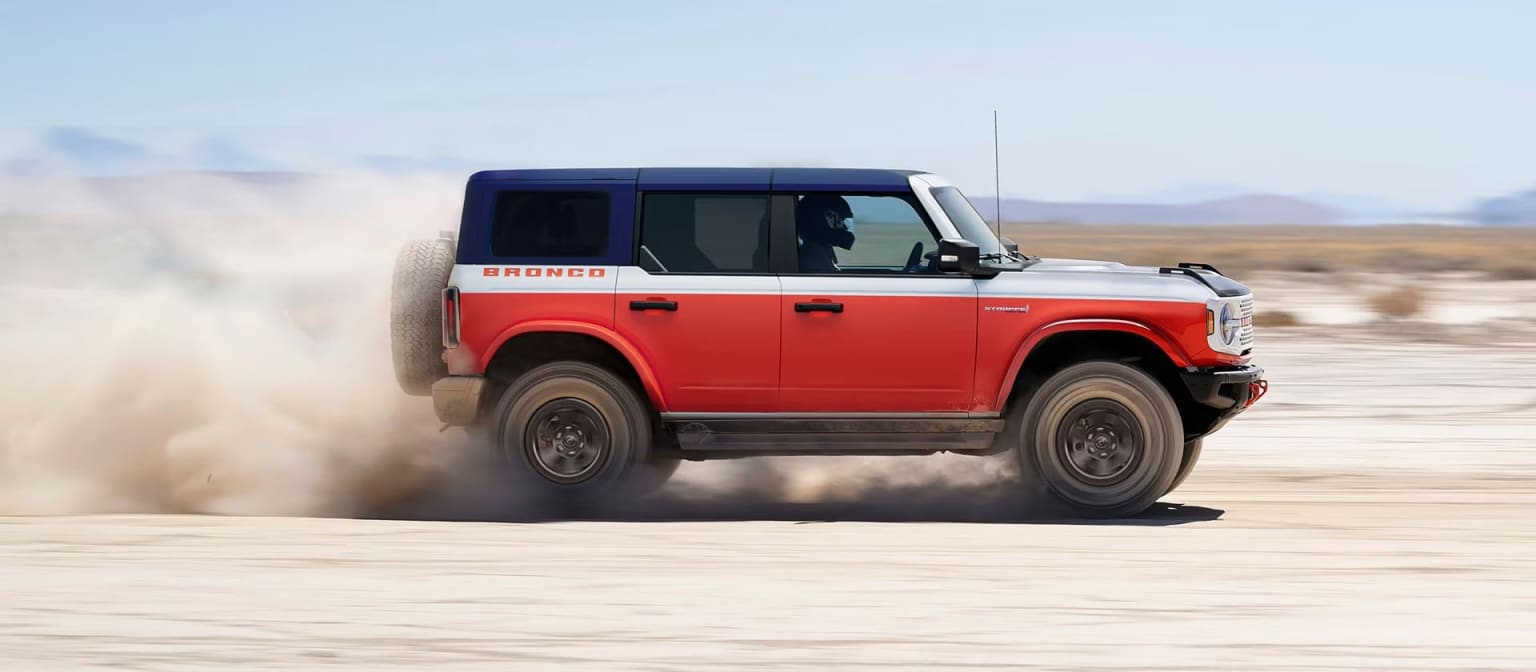 A red, white, and blue SUV kicks up dust as it drives across a dry, desert landscape under a clear sky.