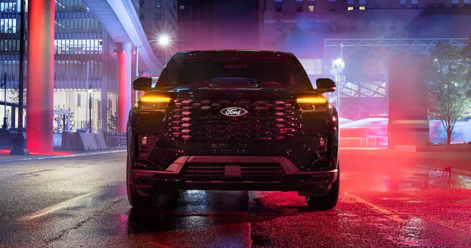 An SUV with glowing headlights parked on a wet street, illuminated by city lights at night.