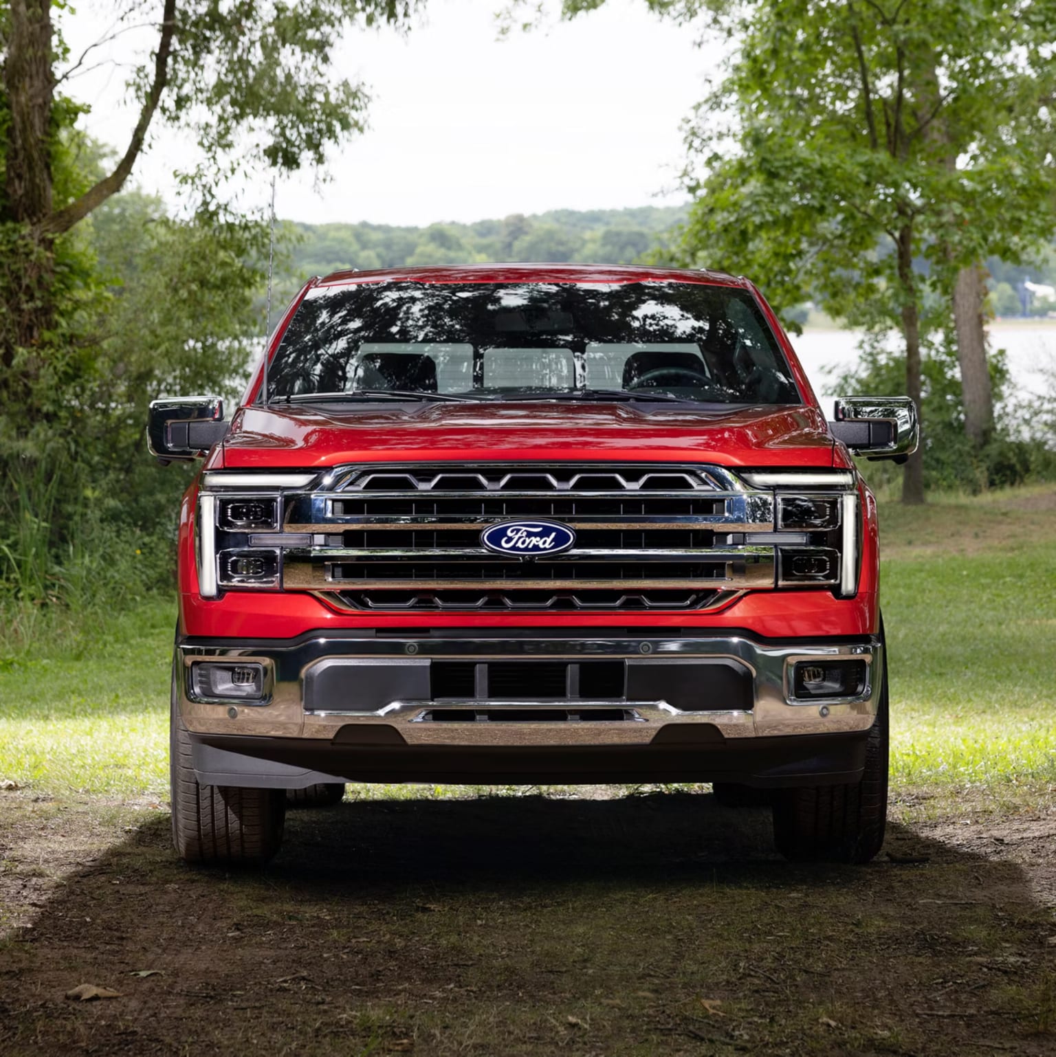 Red pickup truck on dirt path with trees and water in background.