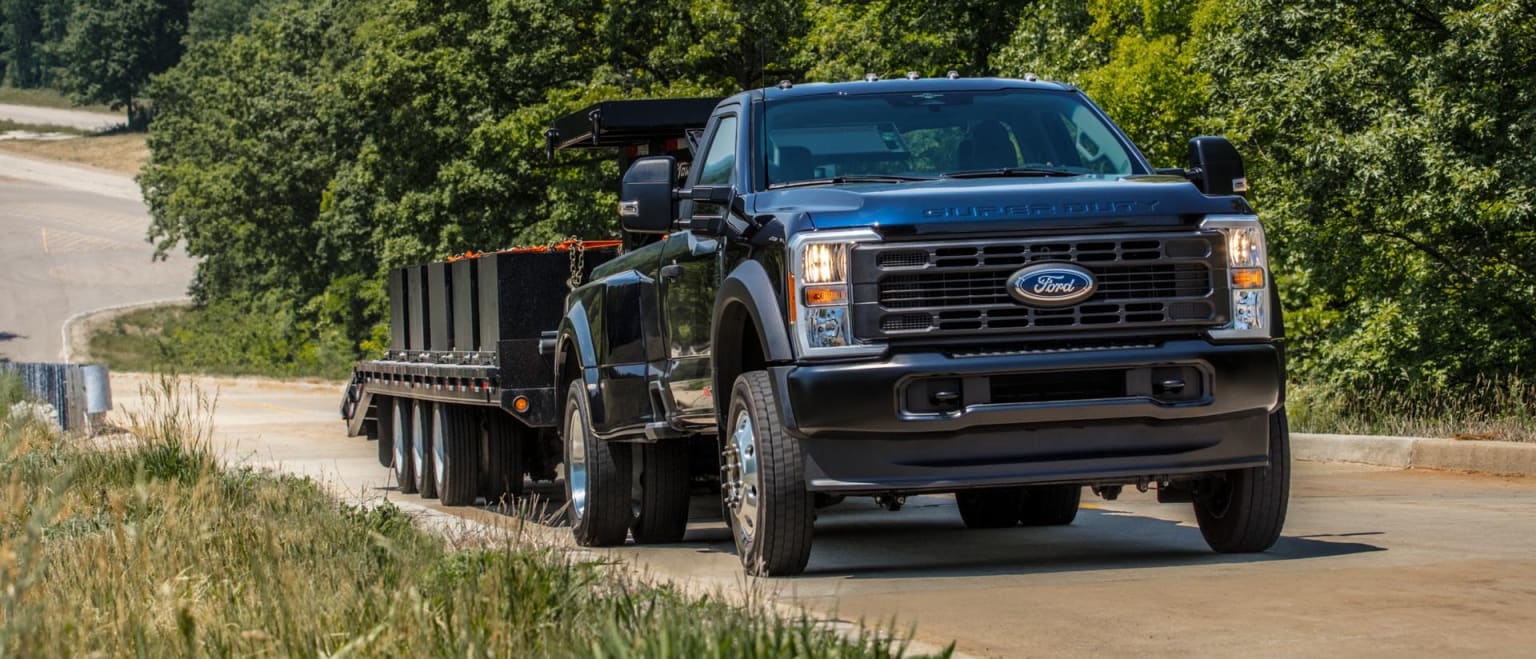 A large black pickup truck with a dump bed attachment is driving on a dirt road surrounded by lush green vegetation.