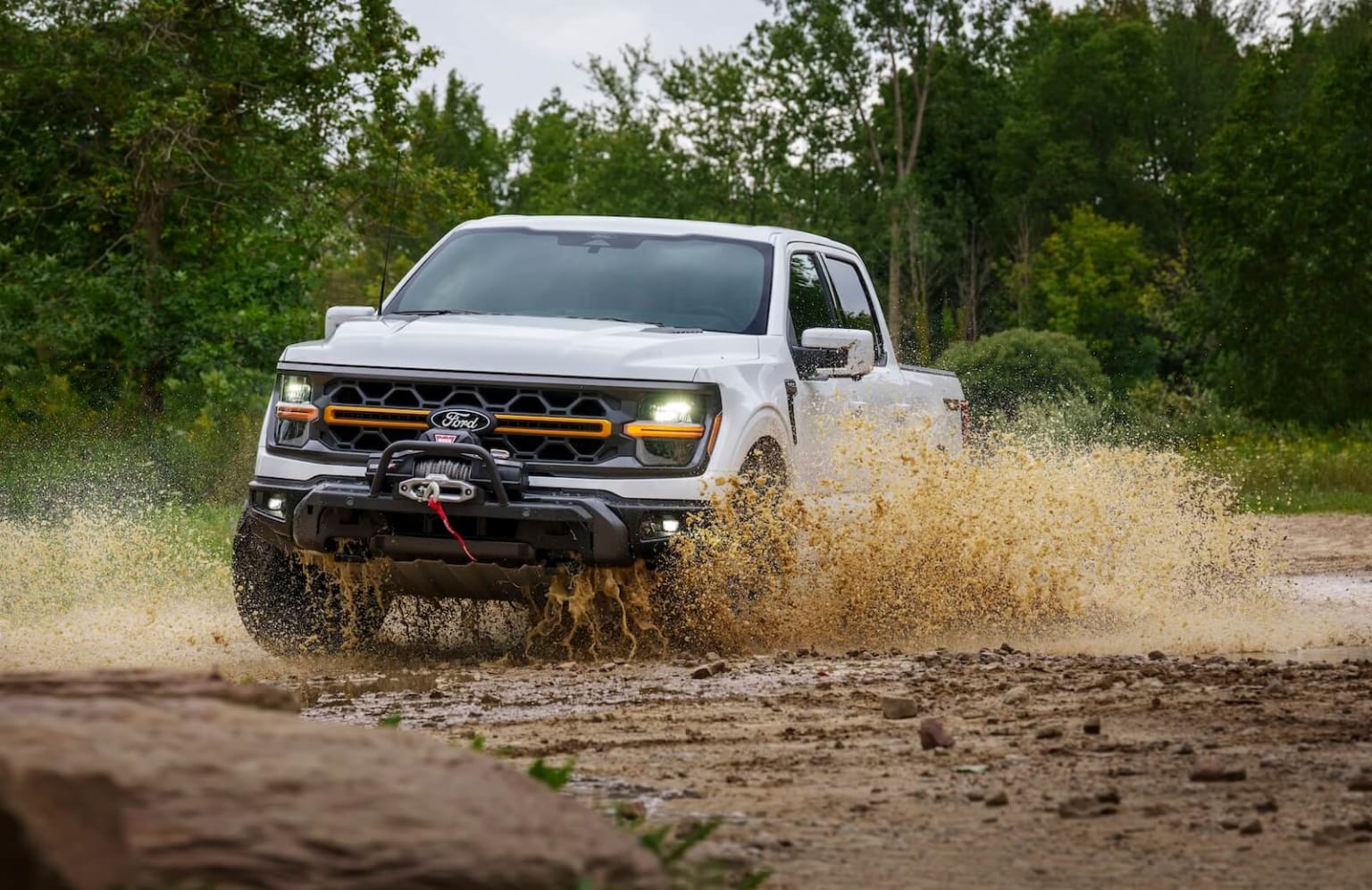 Off-road vehicle driving through muddy dirt road surrounded by lush green trees, kicking up a spray of mud and debris.