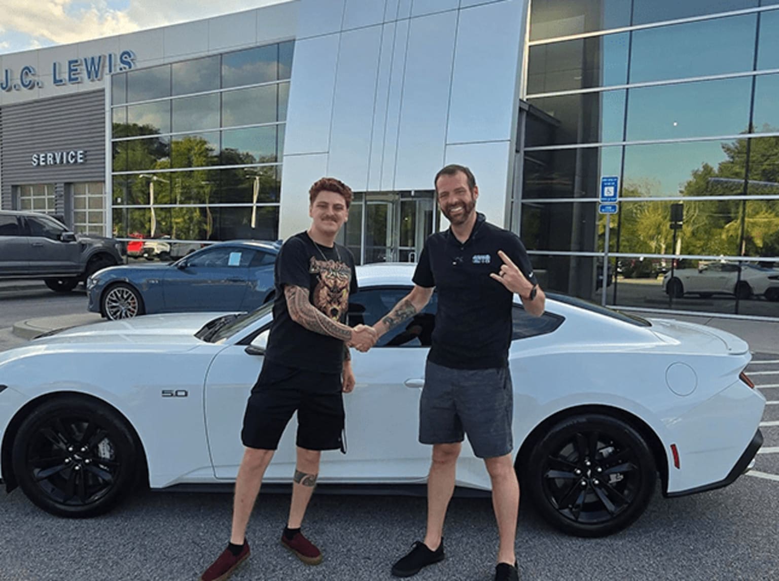 Two men standing in front of a white car parked outside a car dealership.