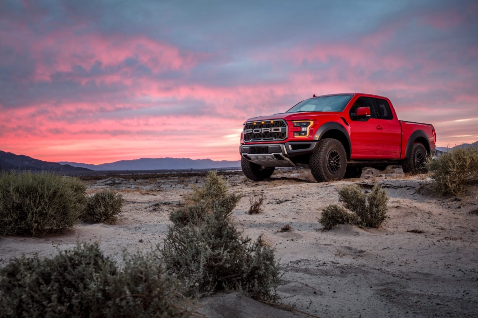 a red truck parked on top of a dirt field next to a field of green grass and bushes at sunset