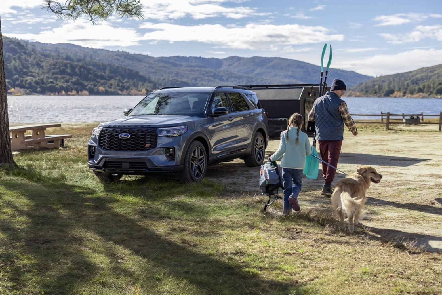 A family walking with their dog near a lake and mountains, with a black SUV parked nearby.