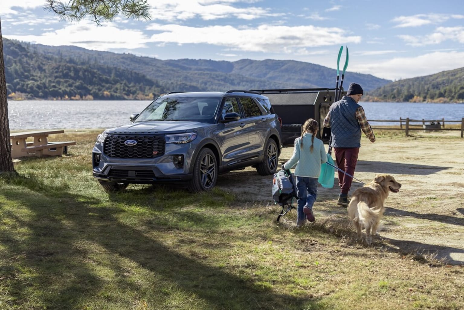 A family walking with their dog near a lake and mountains, with a black SUV parked nearby.