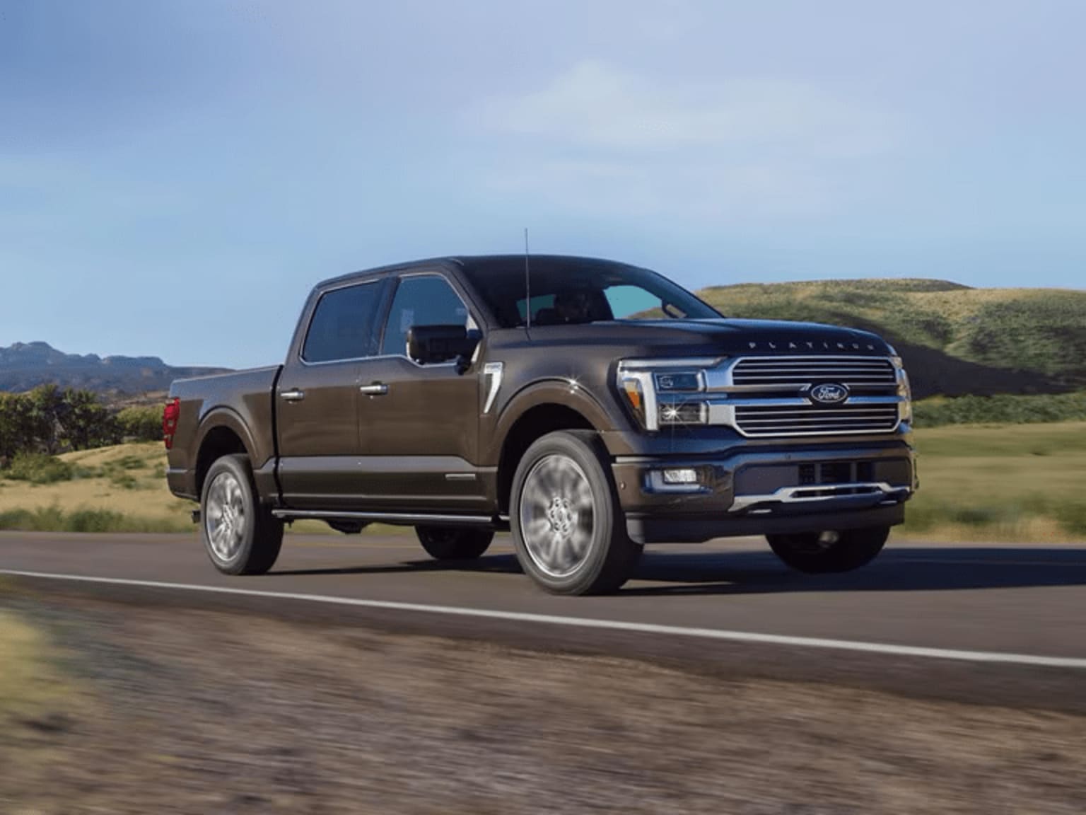 A large, dark-colored pickup truck is driving on a road surrounded by a hilly, grassy landscape under a clear blue sky.
