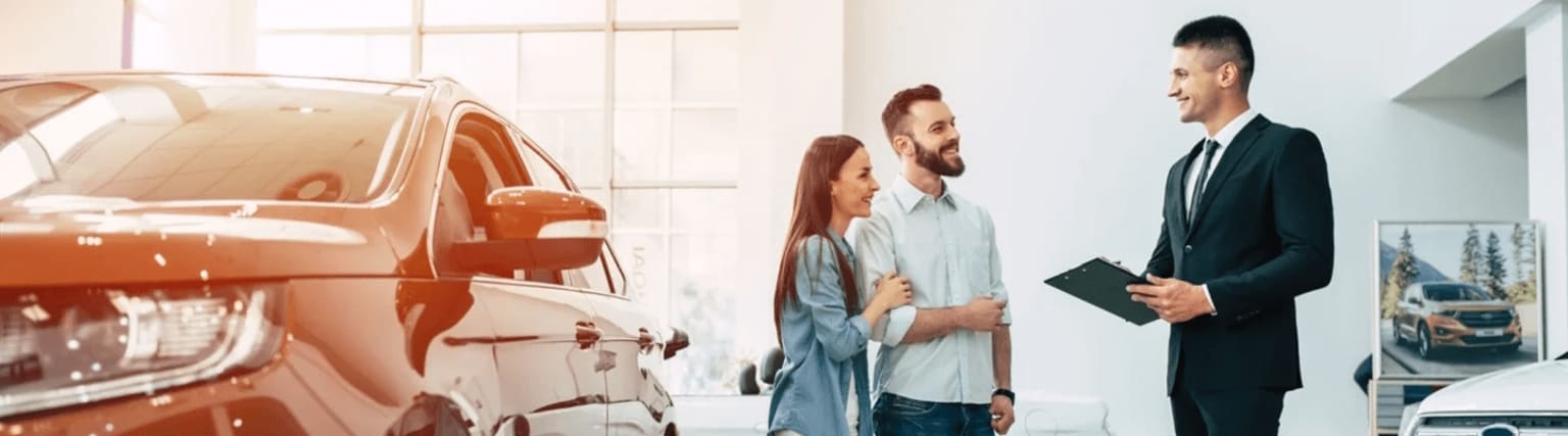 A couple speaking with a man holding a clipboard in a car dealership or showroom.