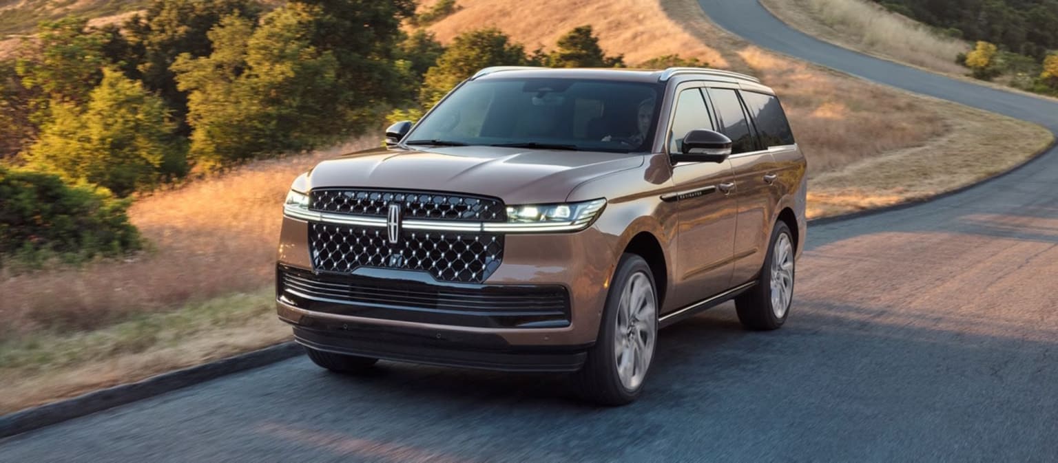 Luxurious SUV in metallic tan color driving down a winding road through scenic hilly landscape with lush forested areas in the background.