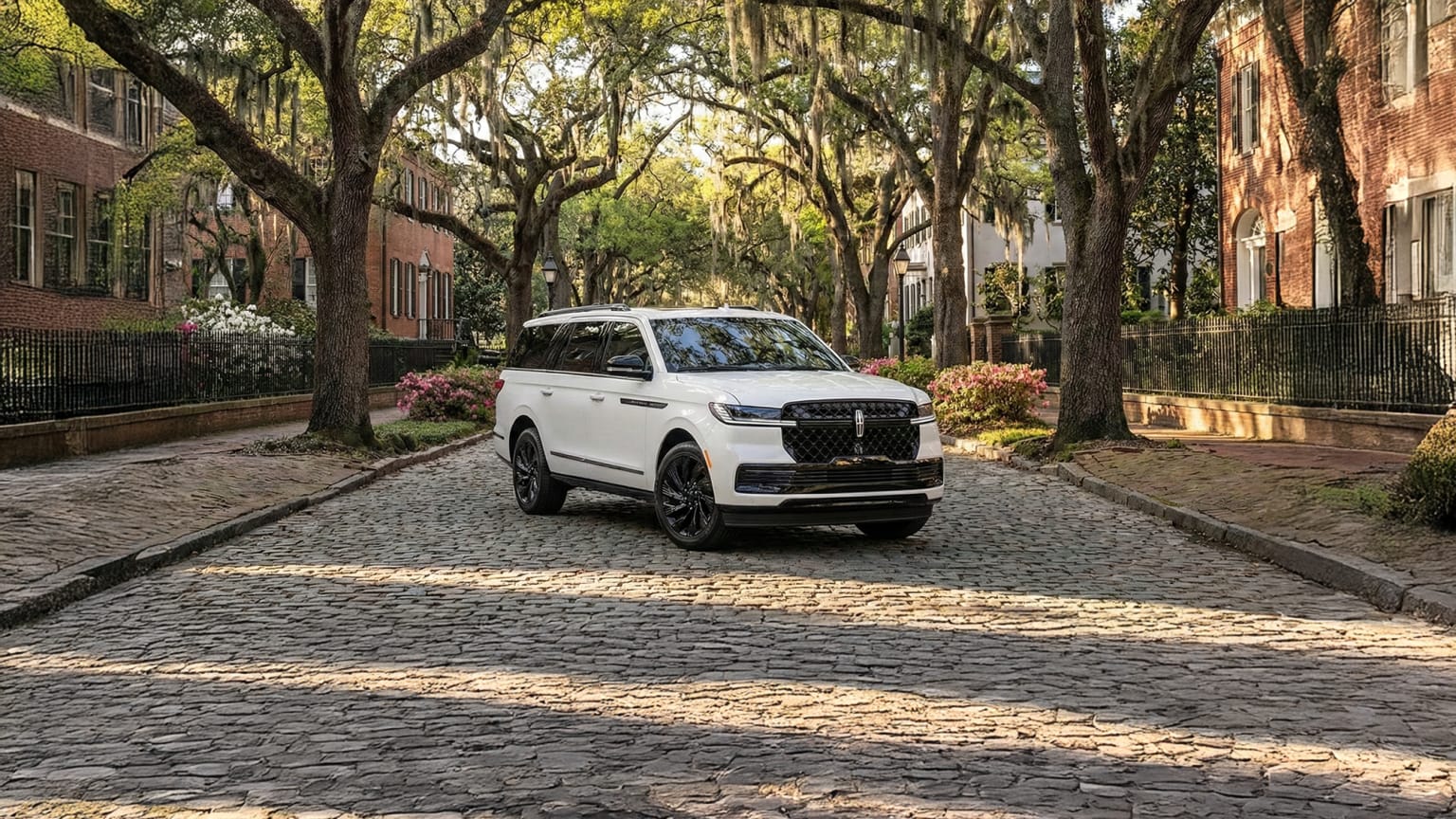 A white SUV parked on a cobblestone street surrounded by lush trees in an urban setting.