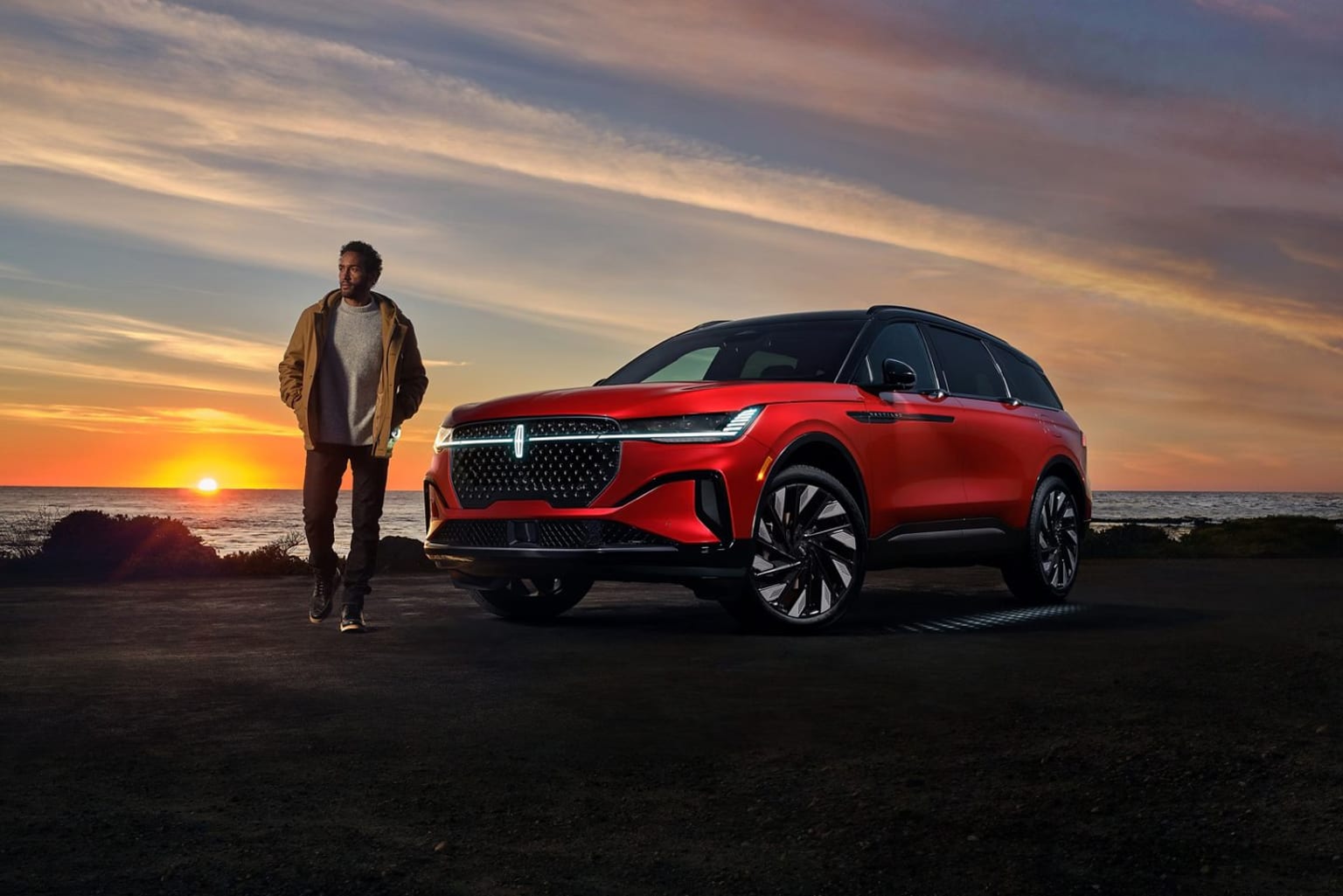 A person stands next to a red SUV against the backdrop of a vibrant sunset over the ocean.