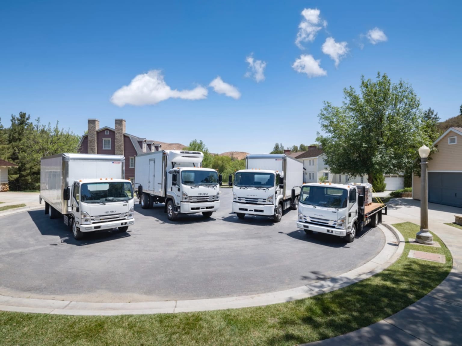 Five trucks parked in a circle on a paved area with houses and trees in the background under a bright blue sky.