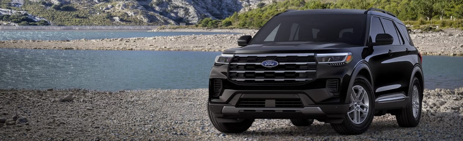 A black SUV is parked on a rocky shore with a calm body of water and mountains in the background.