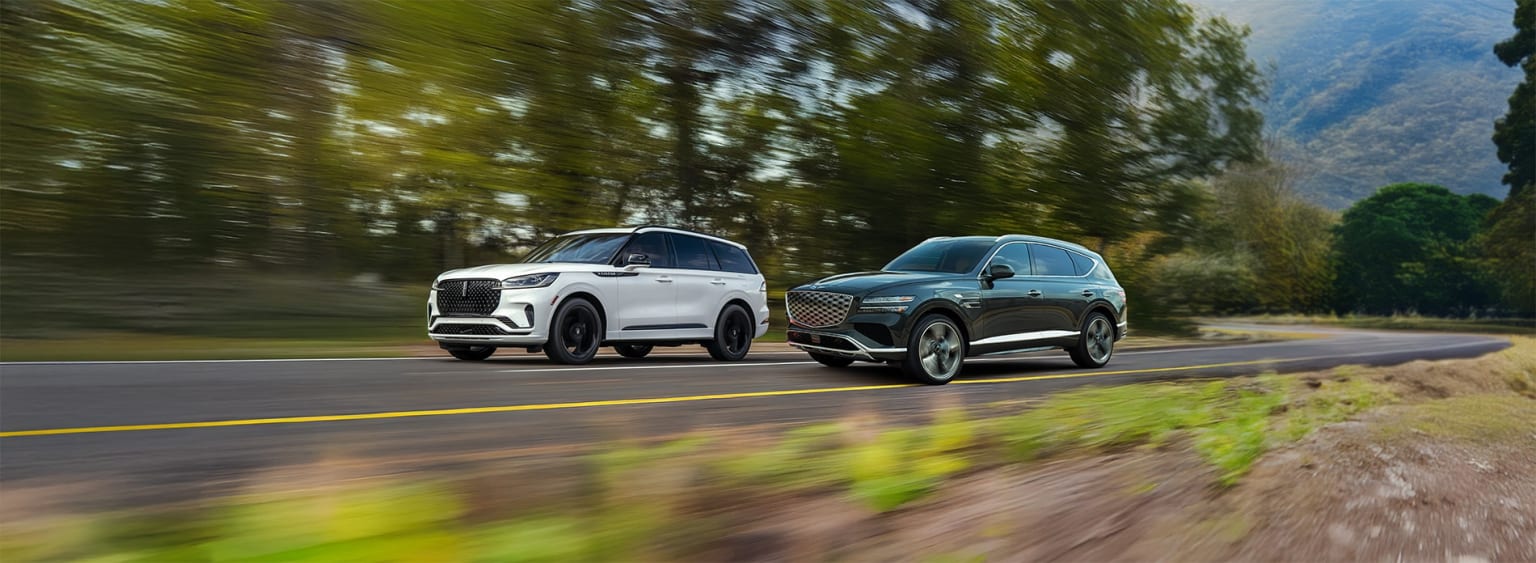 Two large SUVs, one white and one black, are speeding down a winding road surrounded by lush, blurred greenery in the background.