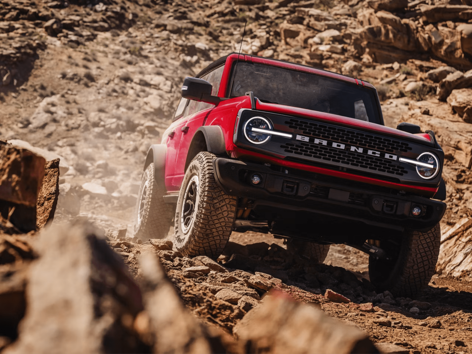 A red SUV navigates a rocky, dusty trail in a desert landscape.