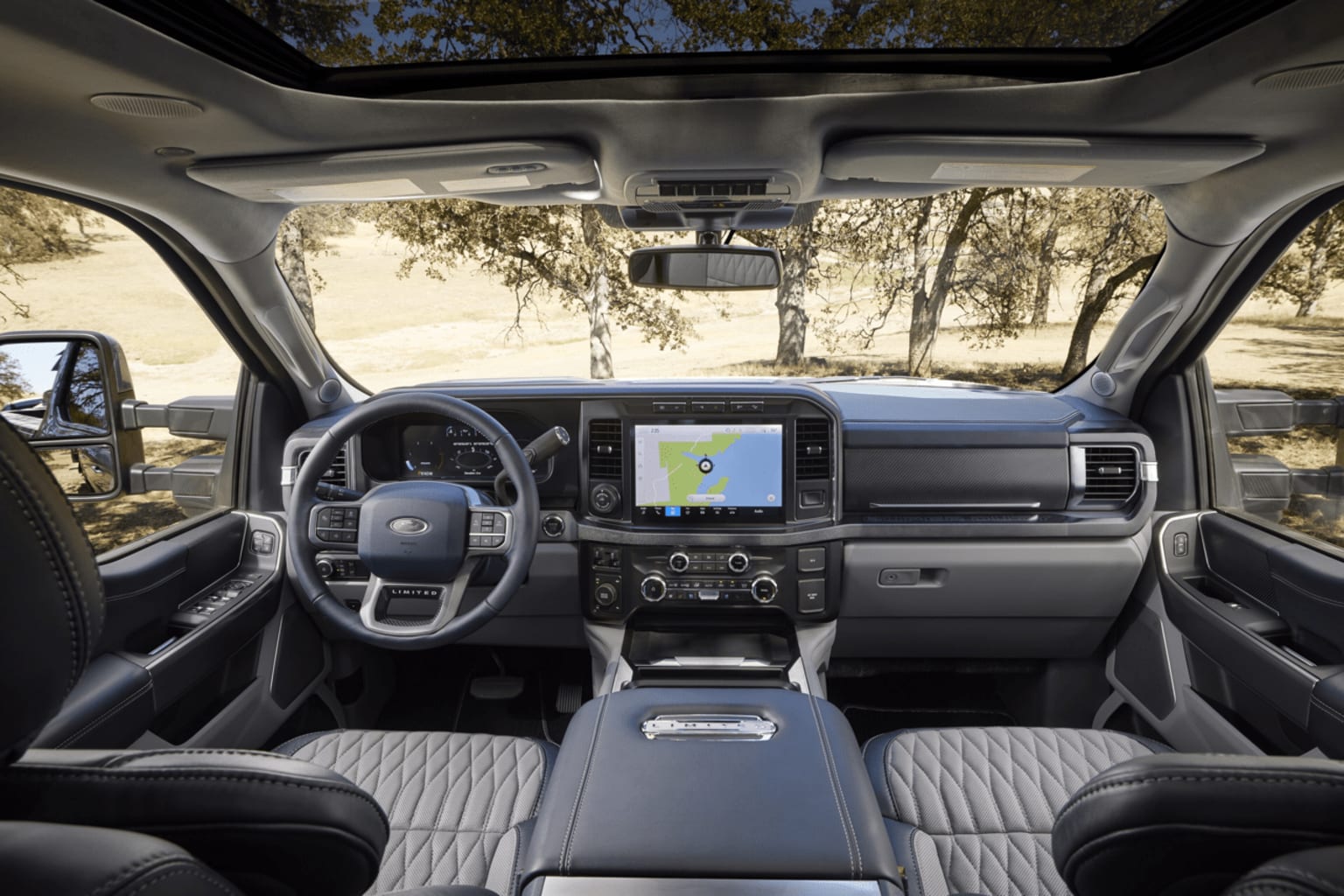 Interior of a modern vehicle with a dashboard featuring a large touchscreen display, steering wheel, and various controls, surrounded by a natural outdoor setting with trees visible through the windshield.