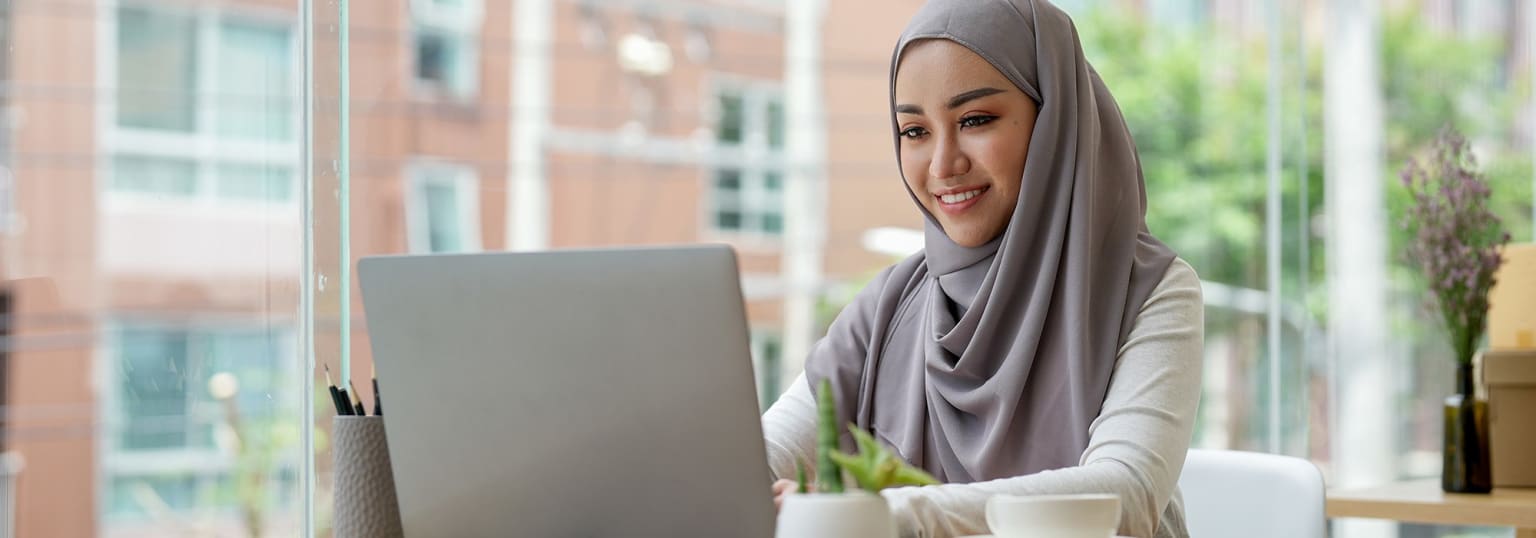 The image shows a young woman wearing a gray hijab, sitting in front of a laptop and smiling at the camera. The background appears to be an indoor setting with a window overlooking a city skyline.