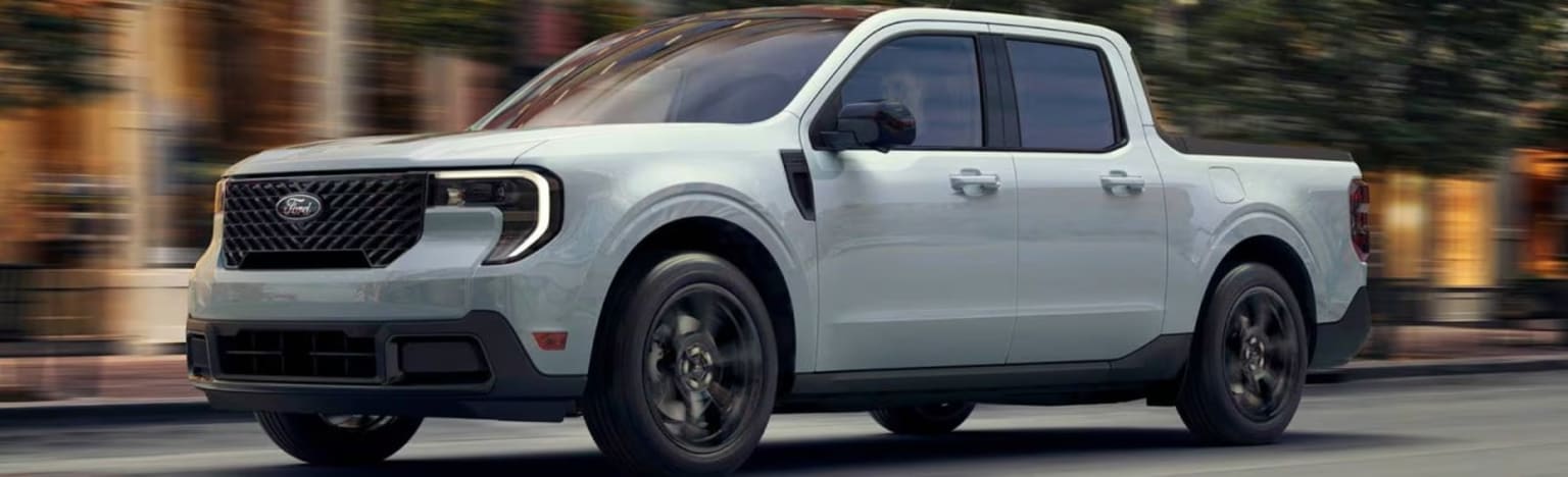 A light-colored pickup truck drives down a blurred street with buildings and trees in the background.