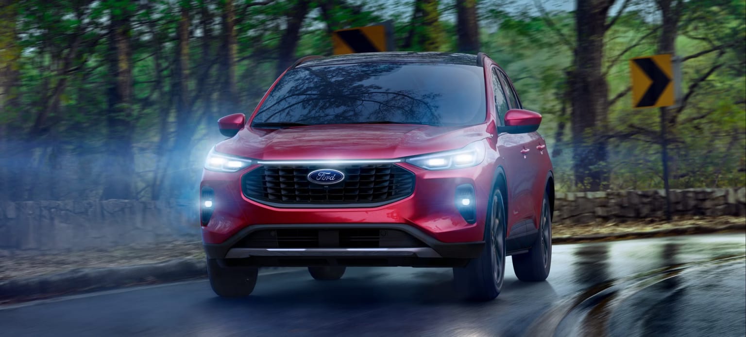 A red compact SUV with bright headlights is driving down a wet, forested road surrounded by tall trees.