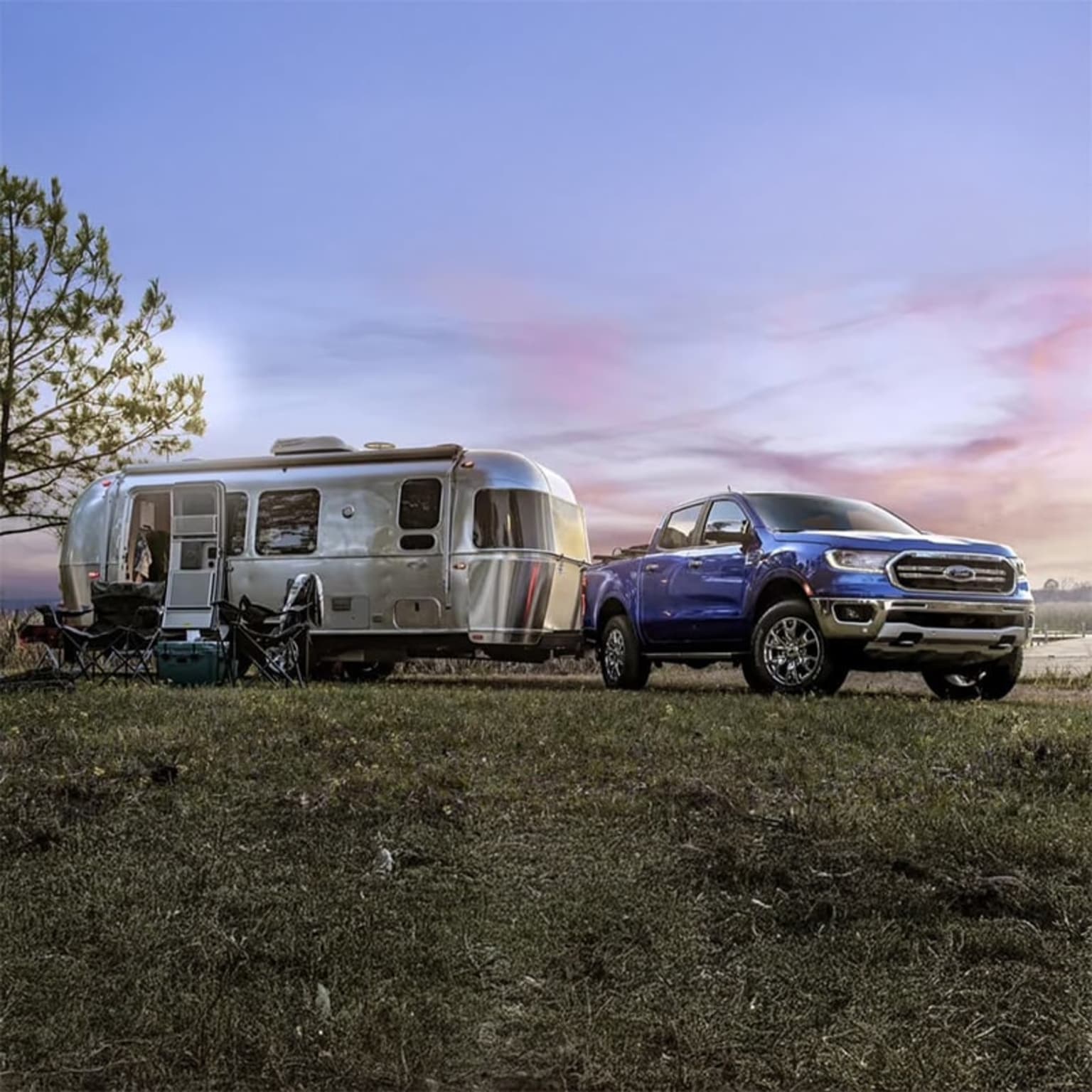 A blue pickup truck is towing a silver Airstream trailer in a grassy field against a backdrop of a colorful sunset sky.
