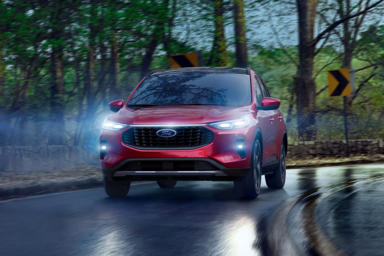 An SUV is driving down a wet road with its headlights on, surrounded by a green forest.