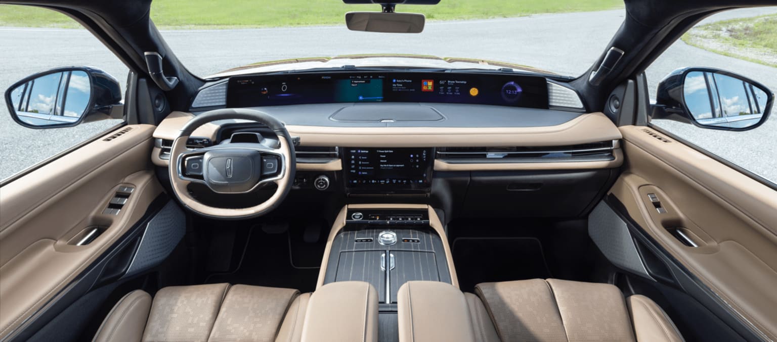 The image shows the interior of a modern, high-tech vehicle with a sleek dashboard, a large central display, and a steering wheel. The background outside the vehicle appears to be a grassy field.