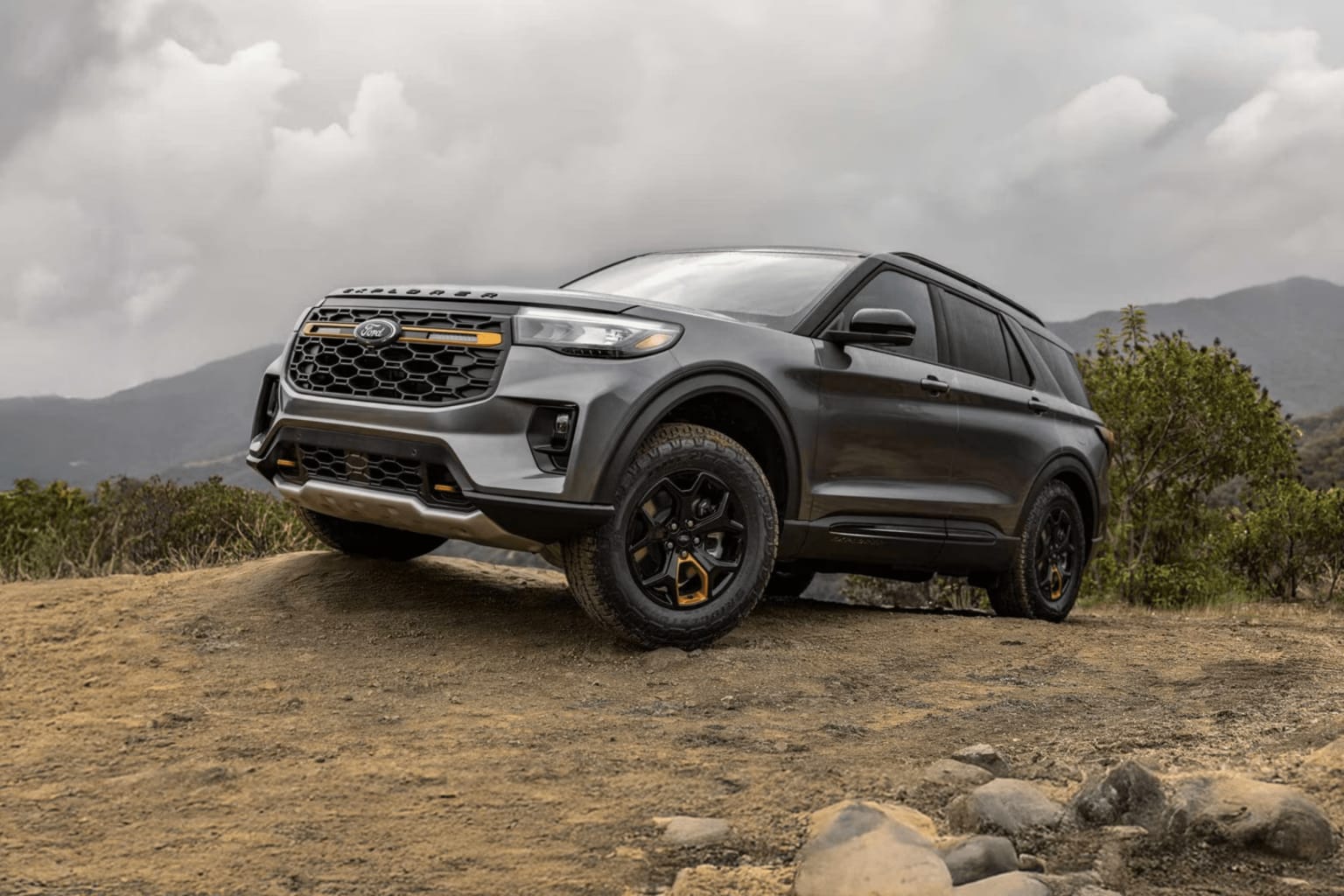 A dark gray SUV with orange accents parked on a dirt hill with rocky terrain in the foreground, with rolling hills and a cloudy sky in the background.