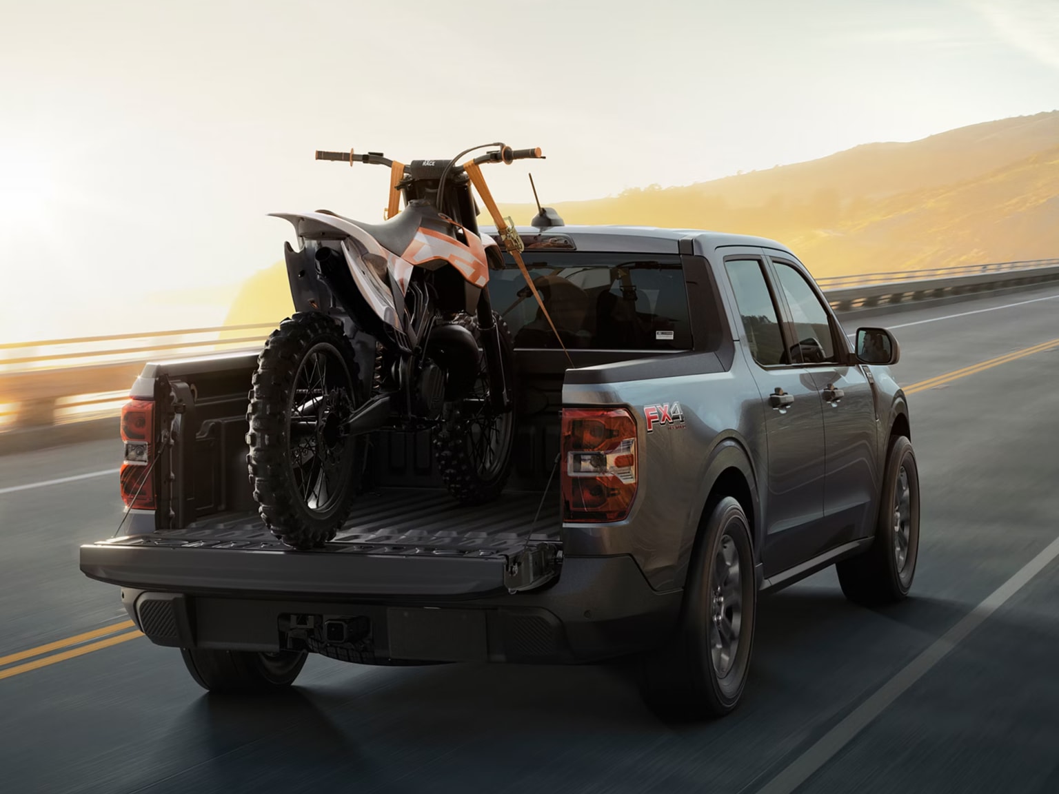 A dirt bike being loaded into the bed of a pickup truck on a road with a hazy, sunlit mountain range in the background.