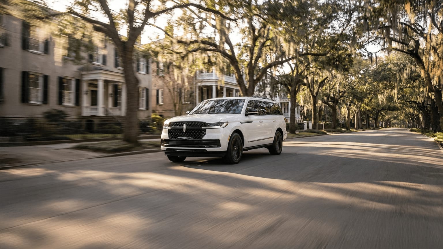 A large white SUV is driving down a tree-lined street, with historic buildings and lush foliage visible in the background.