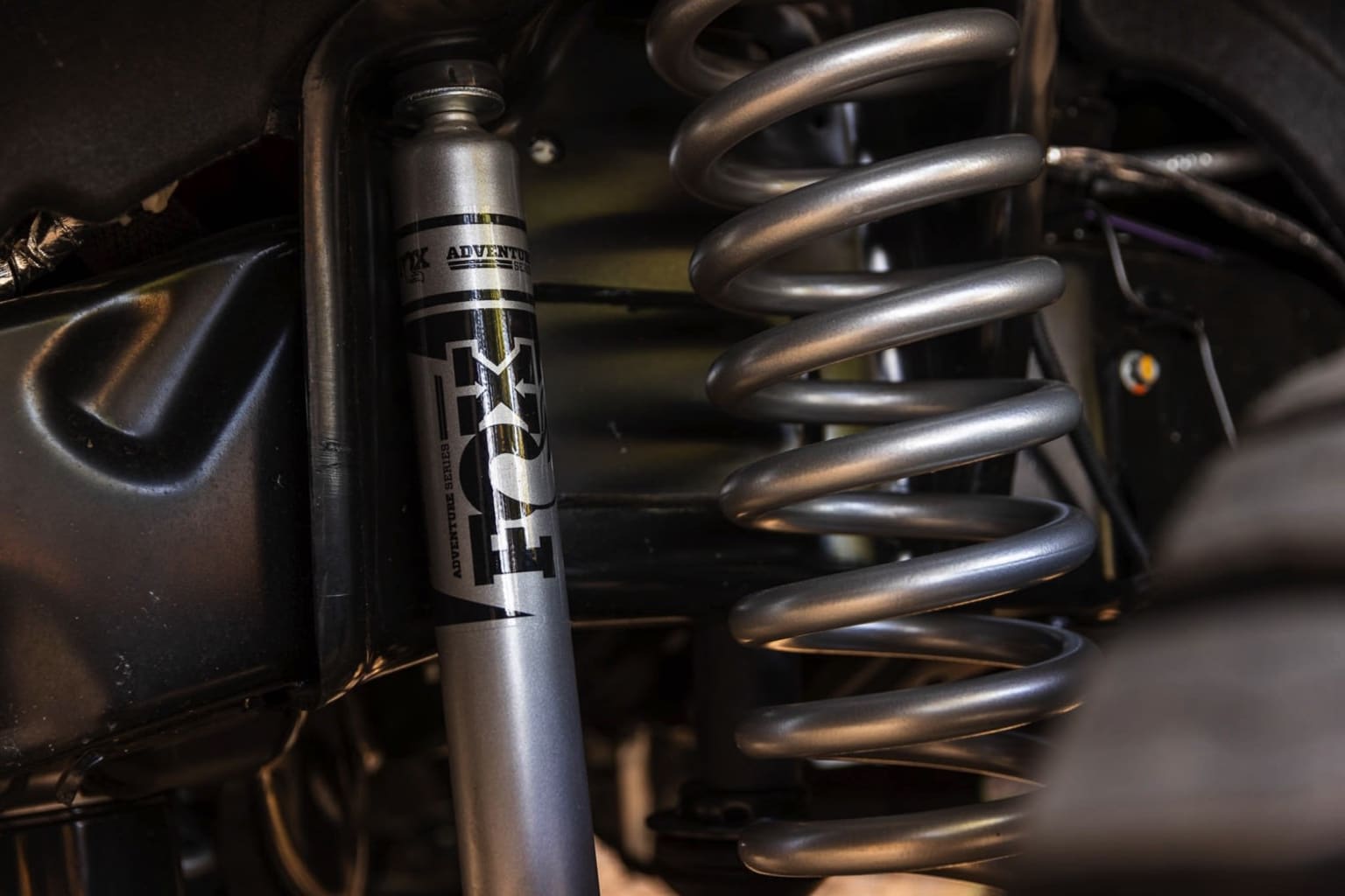 Suspension system of a motorcycle with a silver shock absorber and coiled springs visible in the foreground against a dark background.