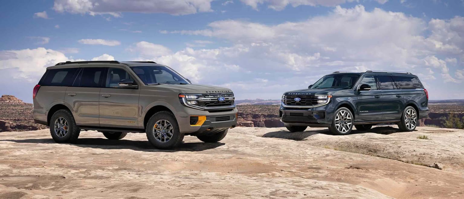 Two SUVs parked on desert landscape with mesas and cloudy sky in the background.
