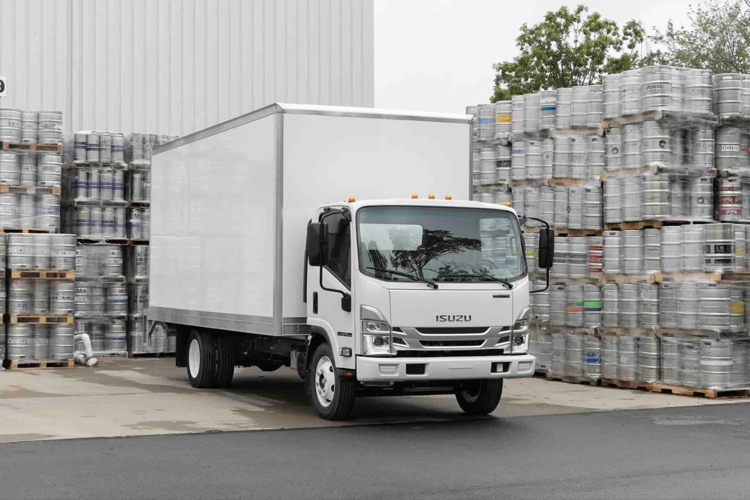 A white box truck parked in front of a large white building with stacks of barrels on either side.